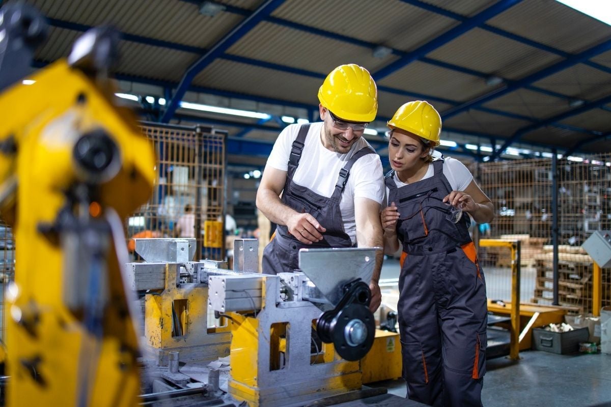 Zwei Fabrikarbeiter mit Schutzhelmen bedienen gelbe Industriemaschine in moderner Produktionshalle – industrielle Fertigung mit Fokus auf Sicherheit und Präzision.