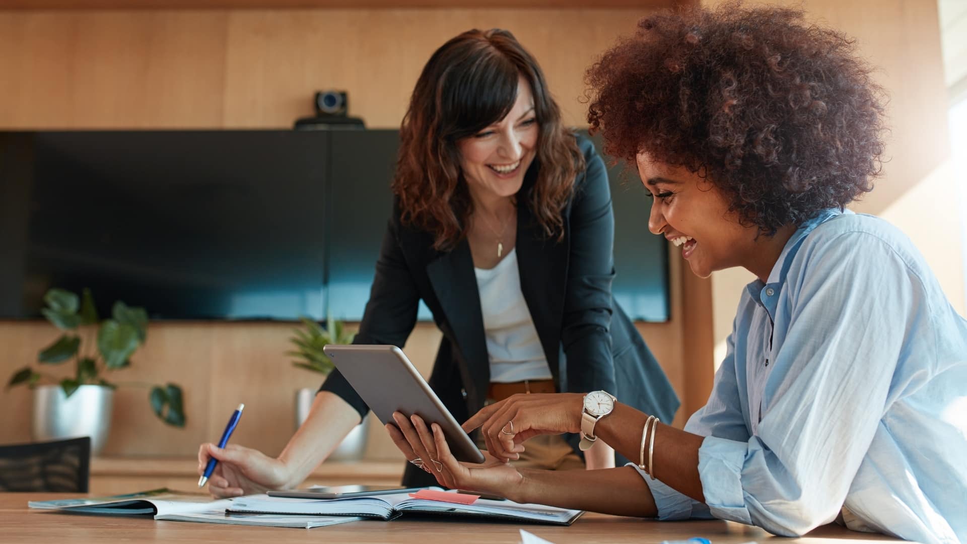 Duas pessoas trabalhando juntas com tablet, caderno e materiais sobre a mesa.