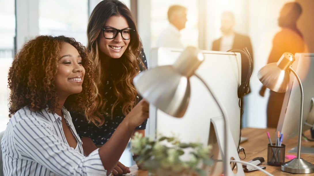 Duas profissionais sorrindo enquanto revisam conteúdo juntas em um computador de mesa, em um ambiente de escritório colaborativo, com colegas ao fundo — representando uma equipe construindo autoridade digital com estratégia de conteúdo bem executada.