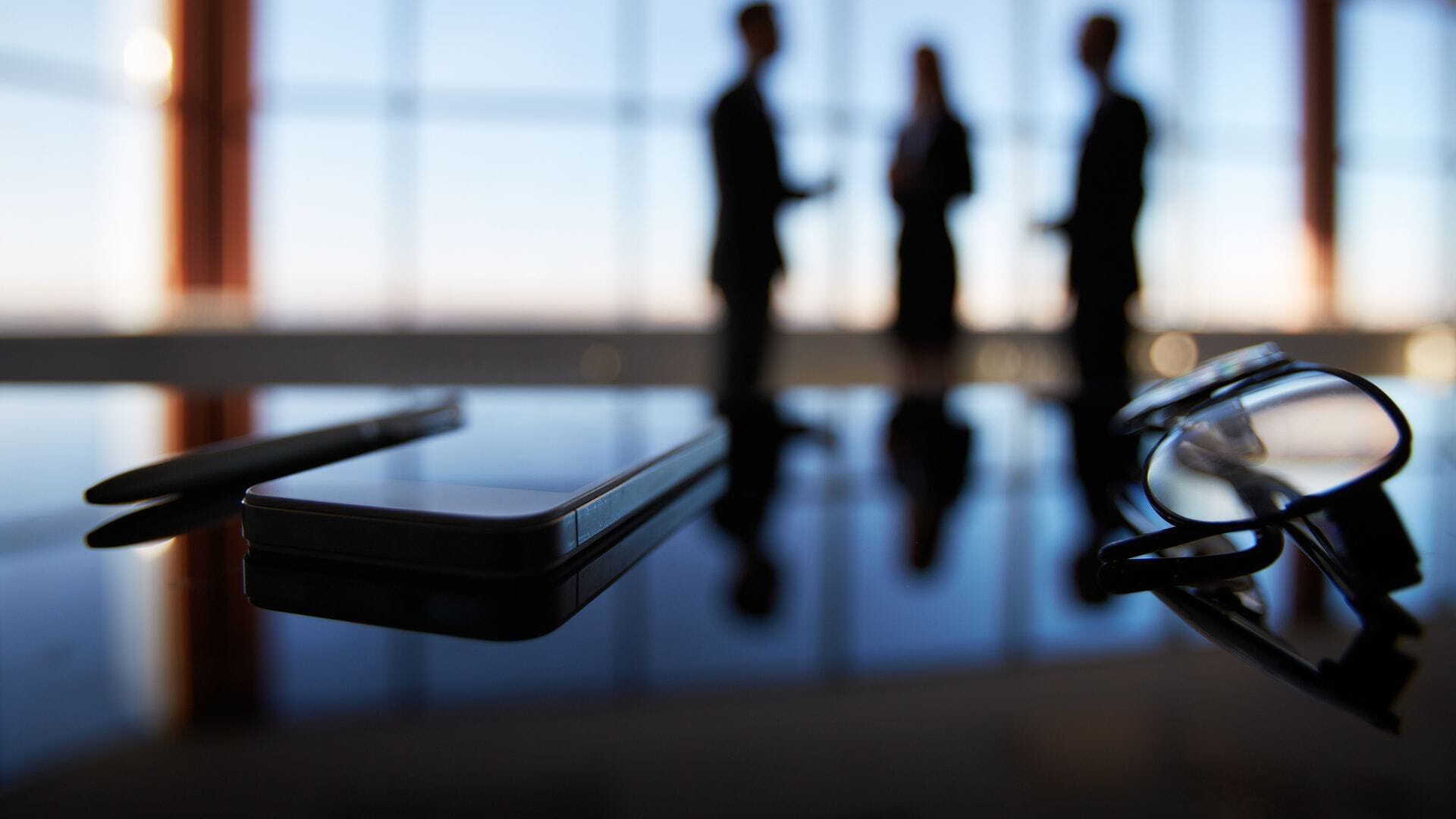 close-up photo of glasses, cell phone, and pen in a work environment, with colleagues interacting in the background